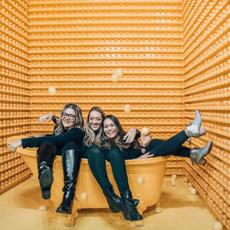 Three women smiling and sitting in a yellow tub surrounded by a wall of rubber ducks, with yellow balls in the air.
