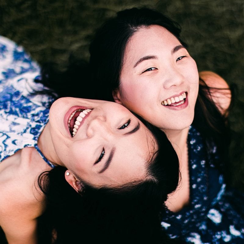Two people lying on grass, smiling and looking upward. Both have dark hair and are wearing patterned tops.