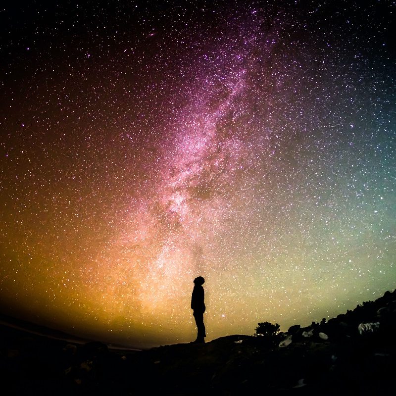 Silhouette of a person standing on a hill, gazing at a sky filled with stars and a bright band of the Milky Way galaxy. The sky displays a gradient of colors from yellow to purple.