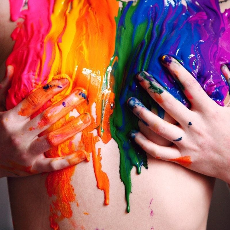 Person's chest covered in vibrant paint colors, including pink, orange, green, and blue. Hands with paint stains rest on the chest, fingers interspersed with the paint.