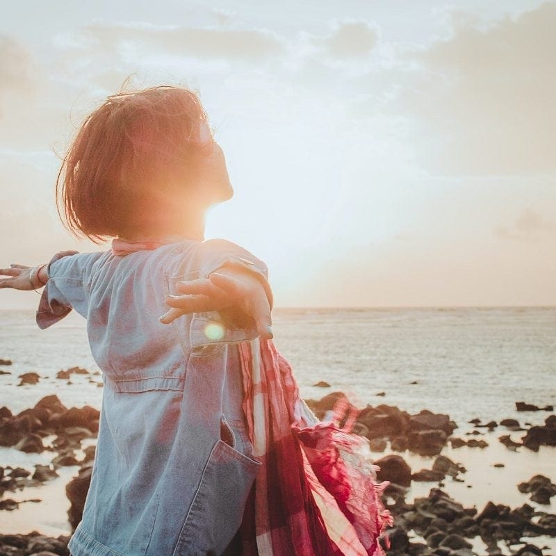 Person with outstretched arms facing the ocean during sunset, wearing a denim jacket. Rocks are visible on the shoreline.