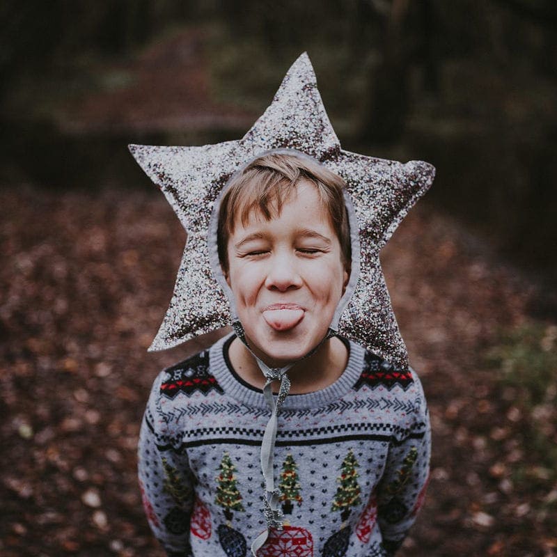 Child wearing a star-shaped hat, sticking out their tongue, stands in a forest wearing a festive sweater.