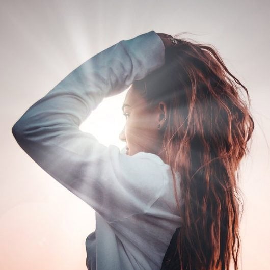 Person with long hair looking towards the sun, hand on head, wearing a white jacket, with visible sunlight rays.