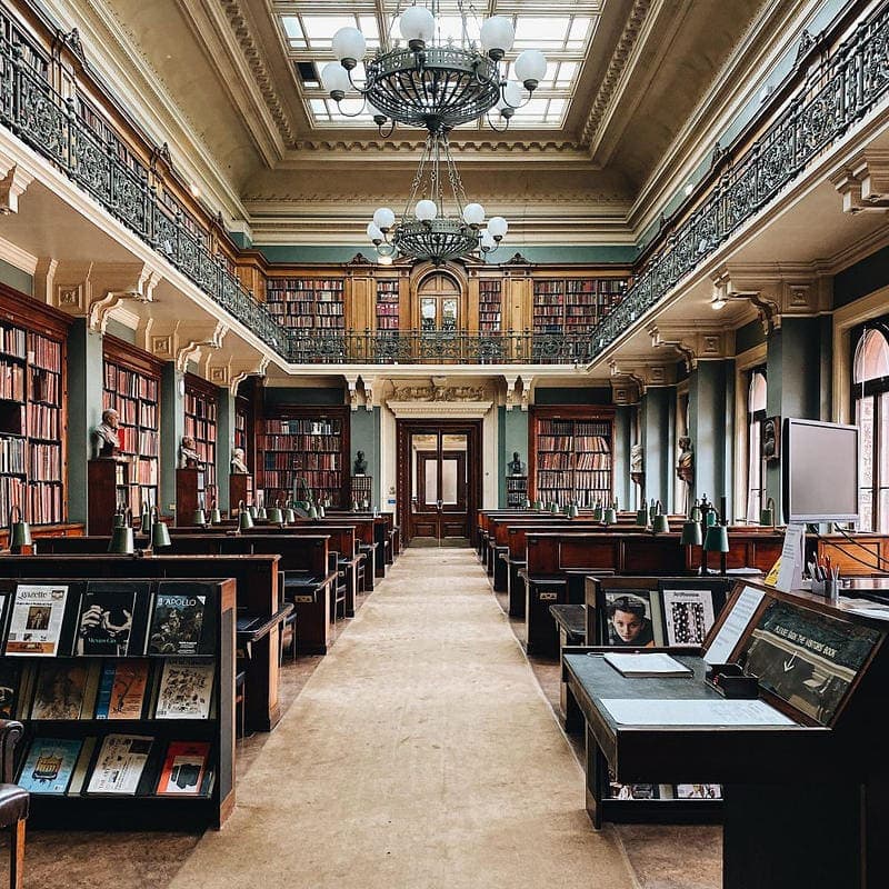 A library interior with wooden furniture, high bookshelves, and a central aisle. Chandelier lights hang from the ceiling, and books and magazines are neatly arranged on tables.